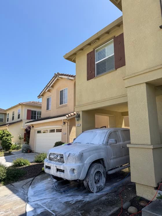 A truck covered in white foam in a residential driveway, mid wash.
